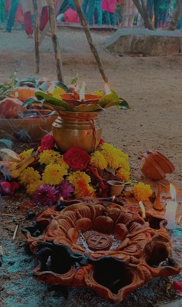 A Chhath arghya setup at dawn — a clay diya thali with lamps, a brass kalash wrapped in kalava and topped with mango leaves, marigold and rose flowers, and a basket of fruit on the ground at the ghat